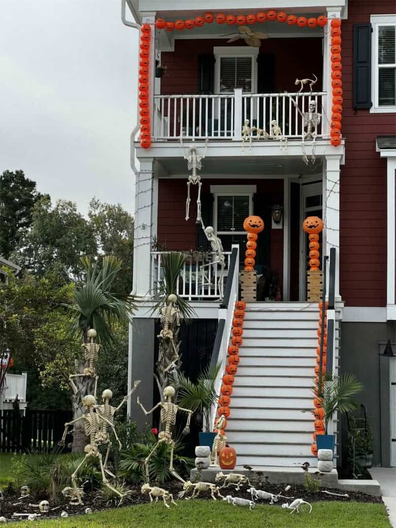 Outdoor Halloween decor features skeletons posed in various ways including climbing trees and hanging on pumpkin lined two story porch.